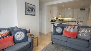 A modern living room with two grey sofas, a small wooden side table, and a grey rug. A kitchen is visible in the background with white cabinets and a black countertop. - Upper Fernlea (Salcombe)