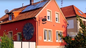 A red building with a large clock, solar panels, and a sign that reads "SPATZEN NEST GASTGEWERK." - Ferienwohnung Spatzennest (Maikammer)
