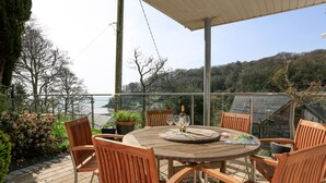 Une table en bois, entourée de quatre chaises, se trouve sur un balcon doté d’une balustrade en verre, d’où l’on peut admirer un plan d’eau et des collines.