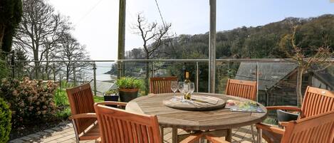Una mesa de madera con cuatro sillas en un balcón con barandilla de cristal, con vistas a un cuerpo de agua y colinas.