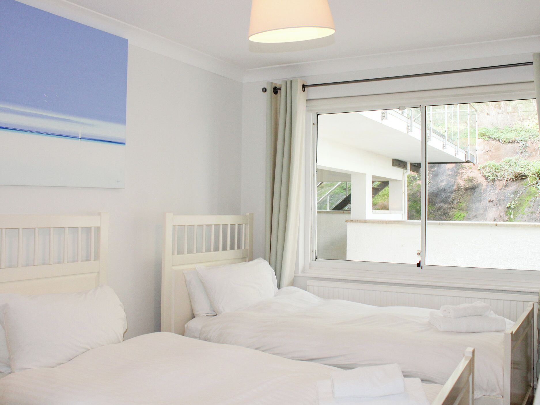 Ein Hotelzimmer mit zwei Betten, einem großen Fenster mit Blick auf ein Gebäude und Grünflächen sowie einer Deckenleuchte.