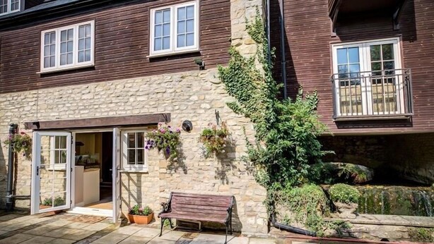 A stone building with a wooden exterior, a bench, and a small garden area. - Mill Race (Burford)