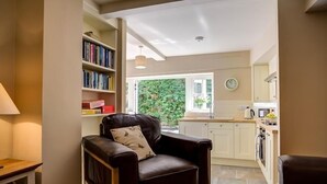 A cozy living room with a leather armchair, a bookshelf, and a view into the kitchen. - Mill Race (Burford)