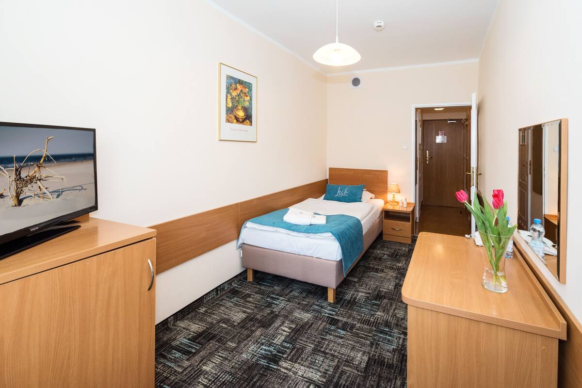 A hotel room with a bed, a desk, a television, a painting, and a vase with flowers.