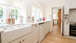 A modern kitchen with white cabinetry, a built-in dishwasher, and a large window with floral curtains. - Home Farm (Witney)