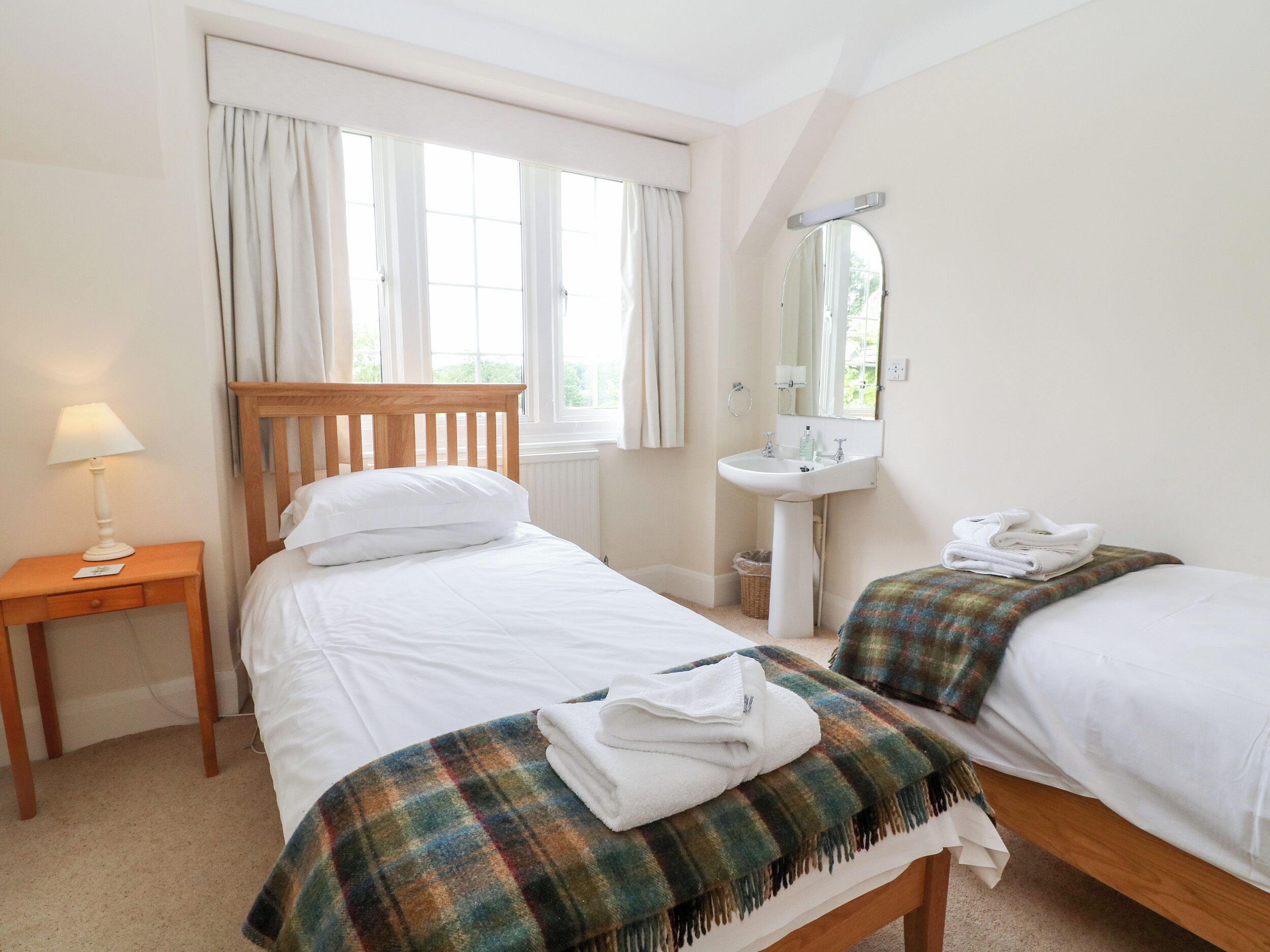 A bedroom with two beds, a bedside table with a lamp, a window with curtains, a sink, and a mirror.