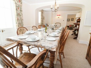Une salle à manger avec une longue table dressée pour six personnes, des chaises en bois, un lustre et une nappe à motifs de cartes géographiques.
