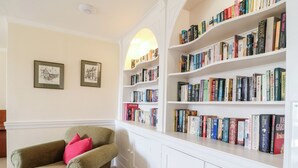 A room with a green armchair, a white bookshelf filled with books, and two framed pictures on the wall. - Home Farm (Witney)