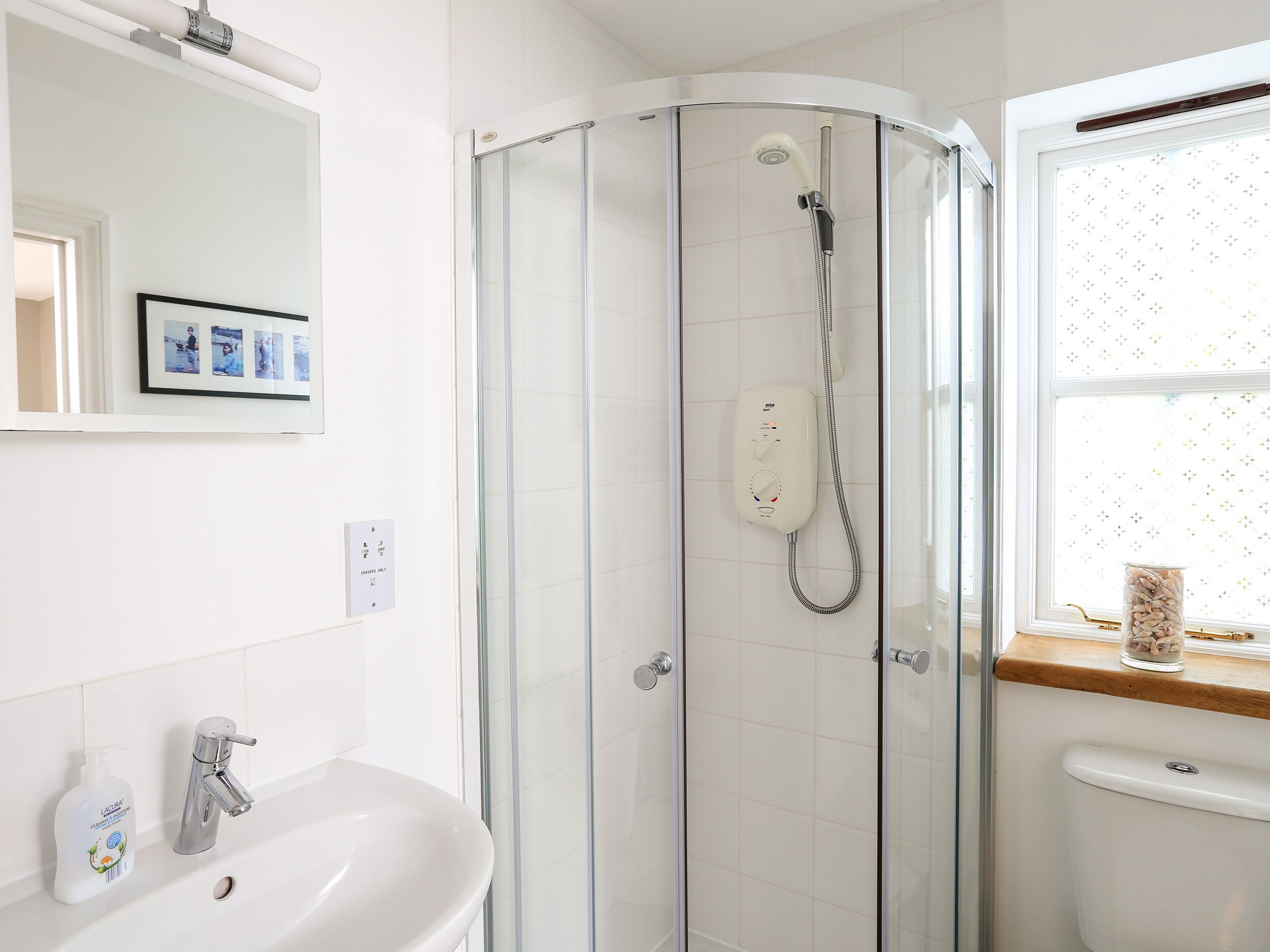 A modern bathroom with a glass-enclosed shower, a white sink with a soap dispenser, and a window with a decorative item on the sill.