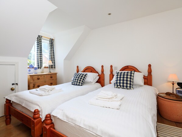 Une chambre avec deux lits en bois, du linge de lit blanc, des oreillers à carreaux, une commode, une lampe et une valise.