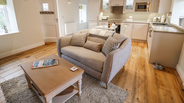 A cozy living room with a sofa, coffee table, and a rug. A kitchen with modern appliances and wooden beams is visible in the background. - The Hayloft (Hawes)