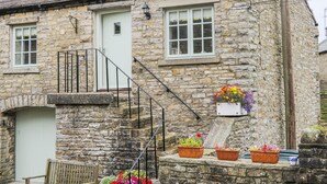 A stone building with a white door, a small porch, and a bench. There are flower pots on the porch. - The Hayloft (Hawes)