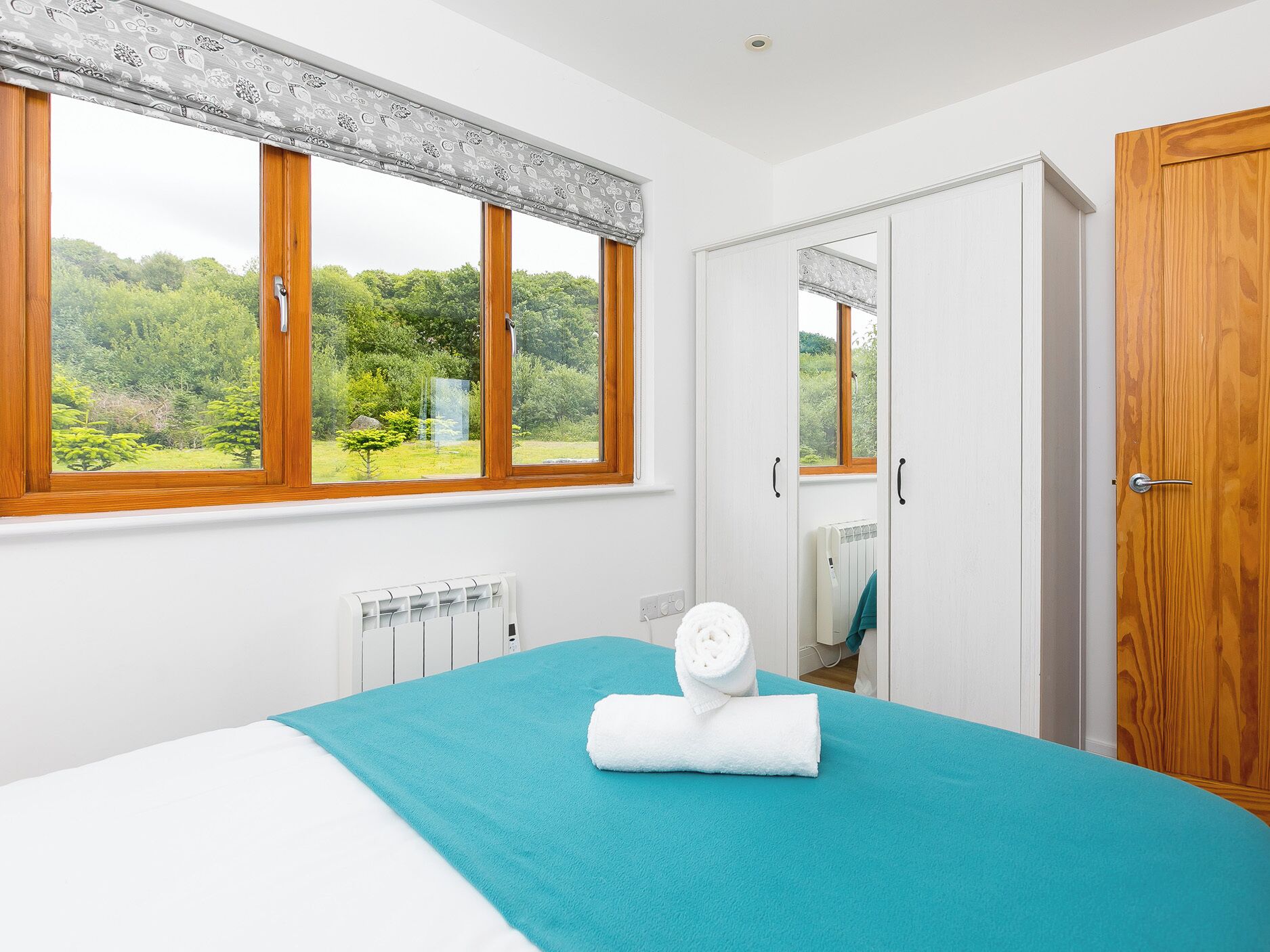 A bedroom with a bed, a window with a view of greenery, a radiator, and a wooden door.