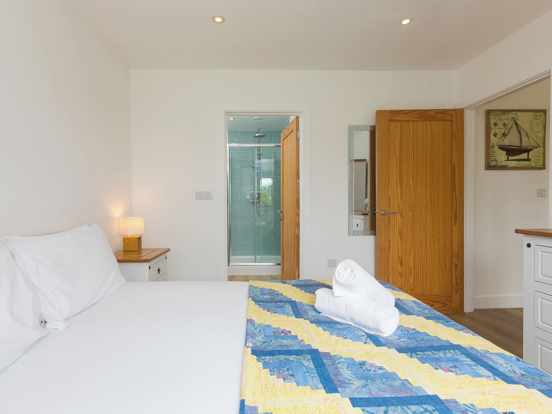 Une chambre d’hôtel avec un lit, une table de chevet avec une lampe, une porte en bois et une salle de bain visible à travers une porte ouverte.