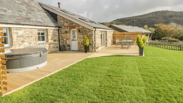 A stone building with a sloped roof, a hot tub, and a patio area with outdoor seating. - Horseshoe Cottage (Pwllheli)
