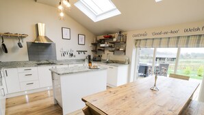 A modern kitchen with white cabinetry, a large island, and a wooden dining table. There are skylights and a view of the outdoors. - Horseshoe Cottage (Pwllheli)