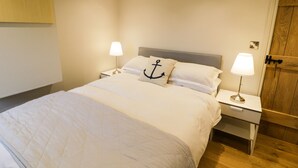 A neatly made bed with a pillow featuring an anchor design, two bedside lamps, and a wooden door in a simple, clean room. - Horseshoe Cottage (Pwllheli)