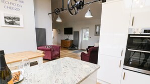 A modern kitchen with white cabinets, a marble countertop, and a built-in oven. There is a dining area with a wooden table and two red sofas. - Horseshoe Cottage (Pwllheli)