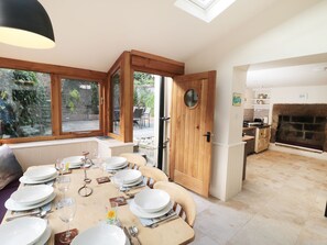 A dining area with a wooden table set for six, a kitchen with a stone fireplace, and a view of the garden through large windows. - Pear Tree House (Whitby)