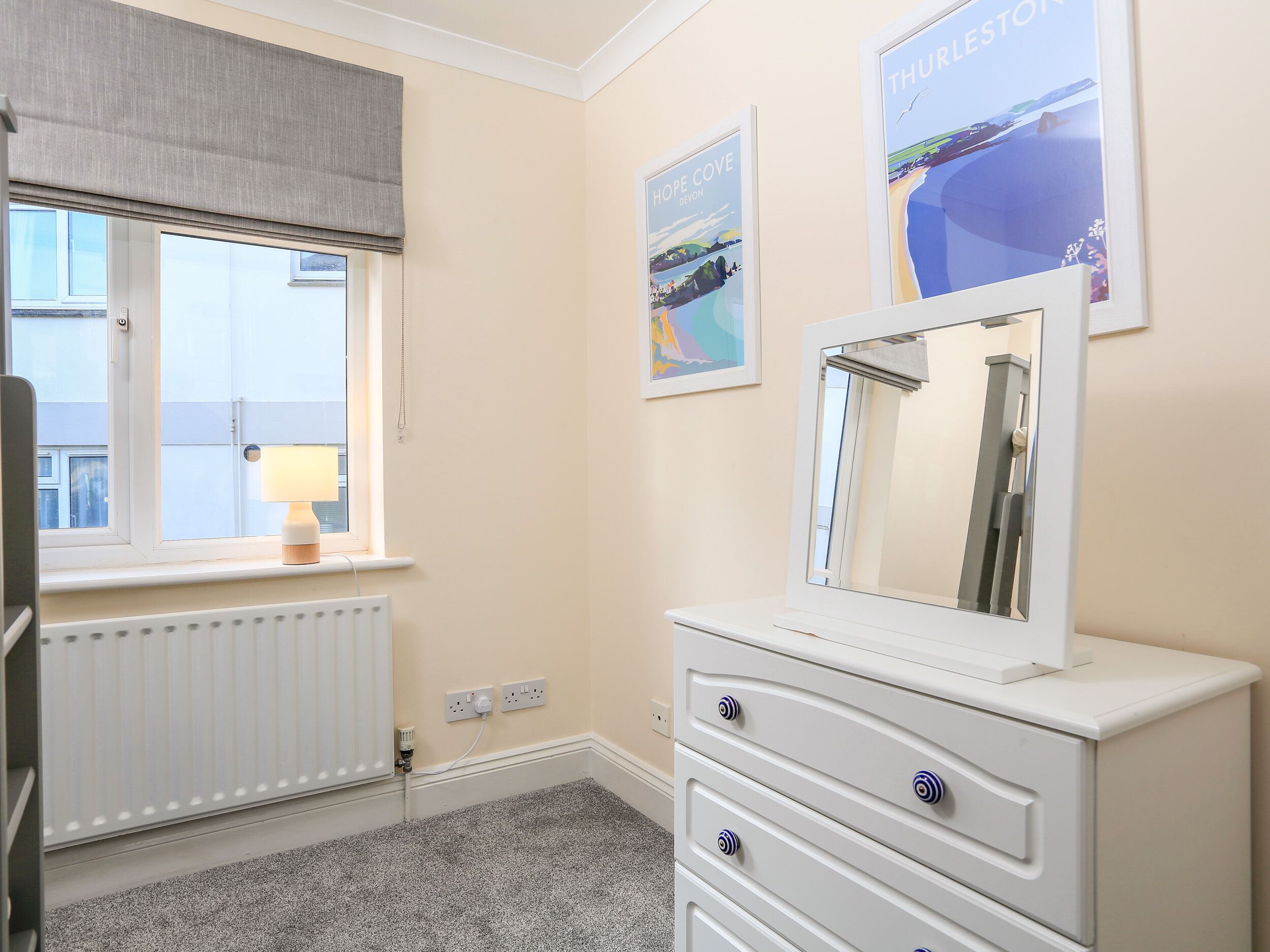 Une chambre avec une fenêtre, un radiateur, une commode blanche et des œuvres d’art accrochées au mur.
