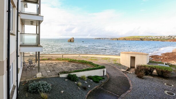A coastal residential area with a view of the sea, a rocky outcrop, and a paved pathway leading to a building with a balcony.
