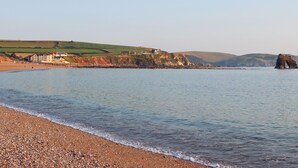 A coastal beach with pebbly shore, a large building with multiple stories, and a prominent rock formation in the water.