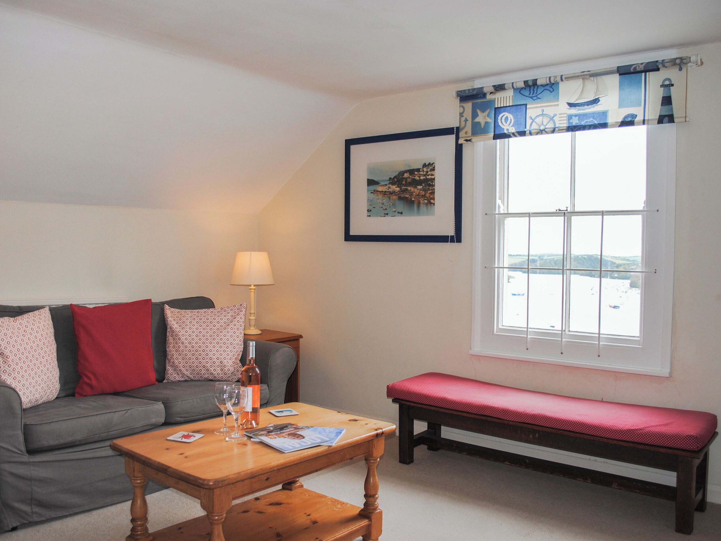 A cozy room with a grey sofa, a wooden coffee table, a red bench, and a window with a view of a body of water.