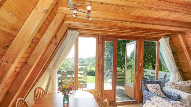 Ein gemütliches, hölzernes Interieur mit einem runden Esstisch, Stühlen und einer Couch mit floralen Kissen.