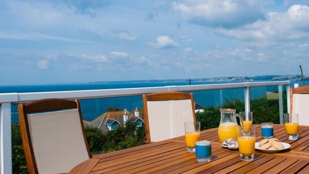 Una mesa de madera con alimentos para el desayuno y vasos de jugo de naranja en un balcón con vista al mar.