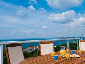 Una mesa de madera con alimentos para el desayuno y vasos de jugo de naranja en un balcón con vista al mar.