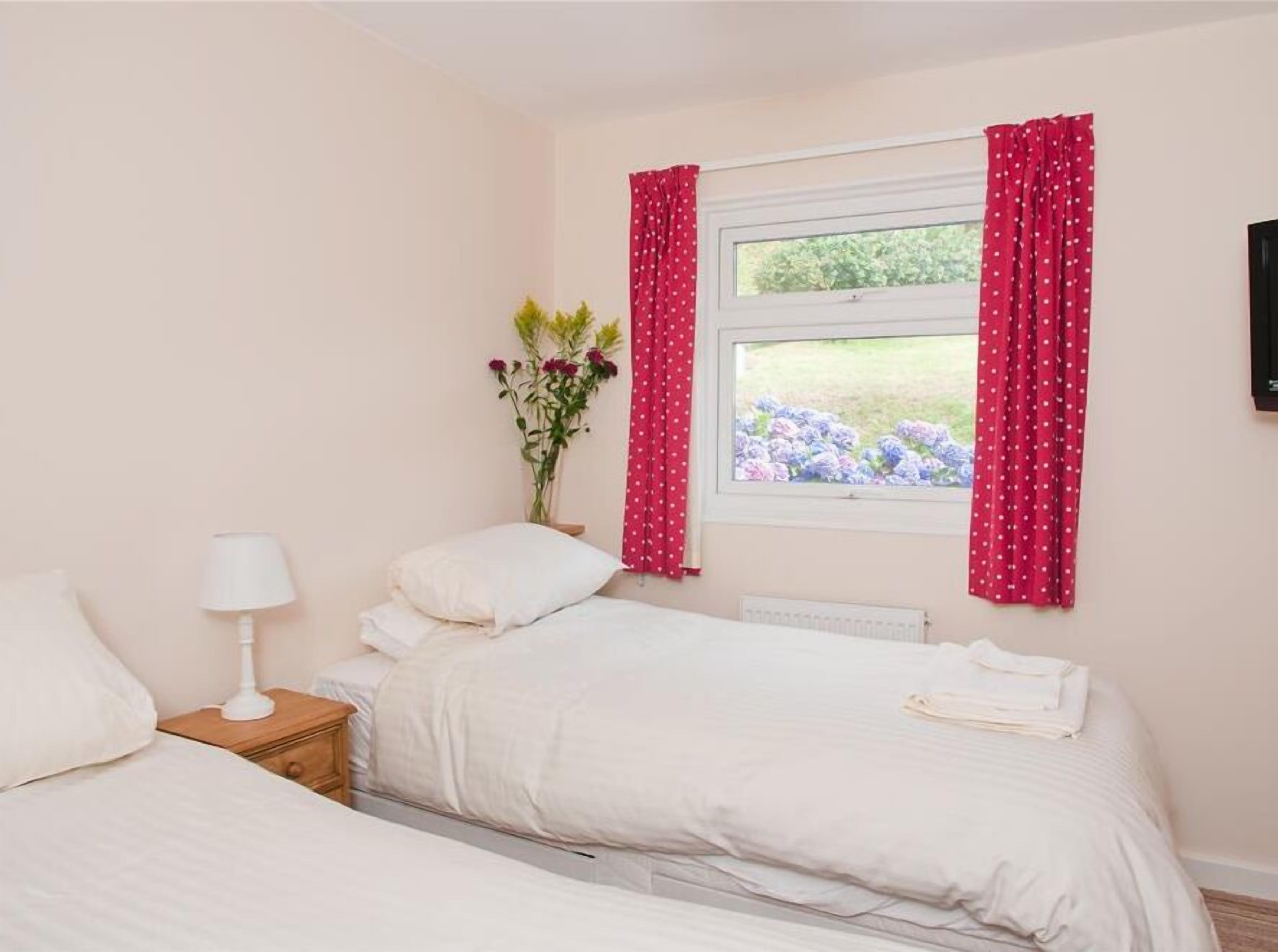 A bedroom with two beds, a nightstand with a lamp, a window with red curtains, and a vase with flowers.