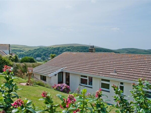 Une maison avec un toit en pente, entourée d’une végétation luxuriante et avec des collines en arrière-plan.