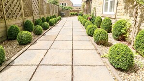 A stone pathway flanked by neatly trimmed hedges and a wooden lattice fence, leading to a traditional stone building with a pitched roof. - South Hill Farmhouse (Cheltenham)