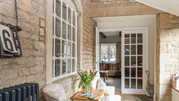 A cozy room with a stone wall, a radiator, a floral armchair, a small table with a vase of flowers, and a view into another room through an open door. - South Hill Farmhouse (Cheltenham)