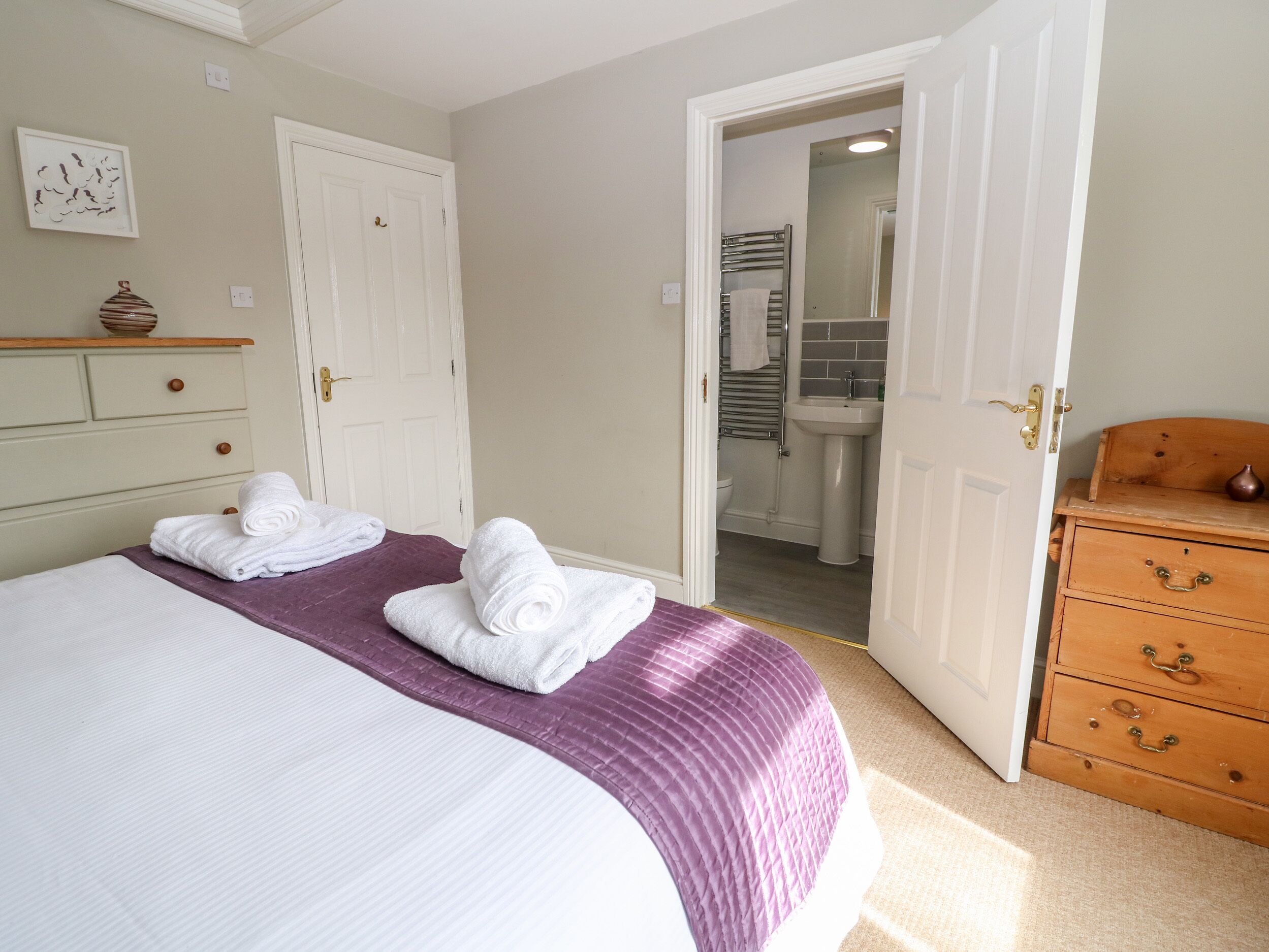 A hotel room with a bed, two rolled towels, a dresser, and a bathroom visible through an open door.