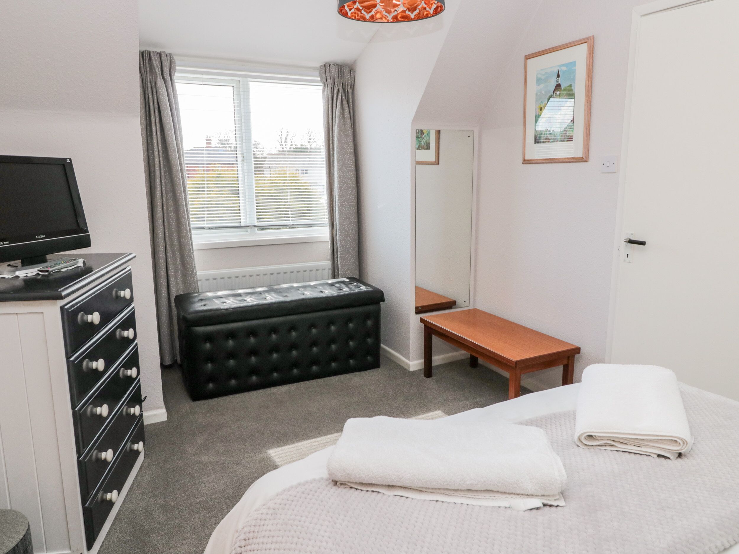 A bedroom with a bed, a desk, a chair, a dresser, a television, and a window with curtains.