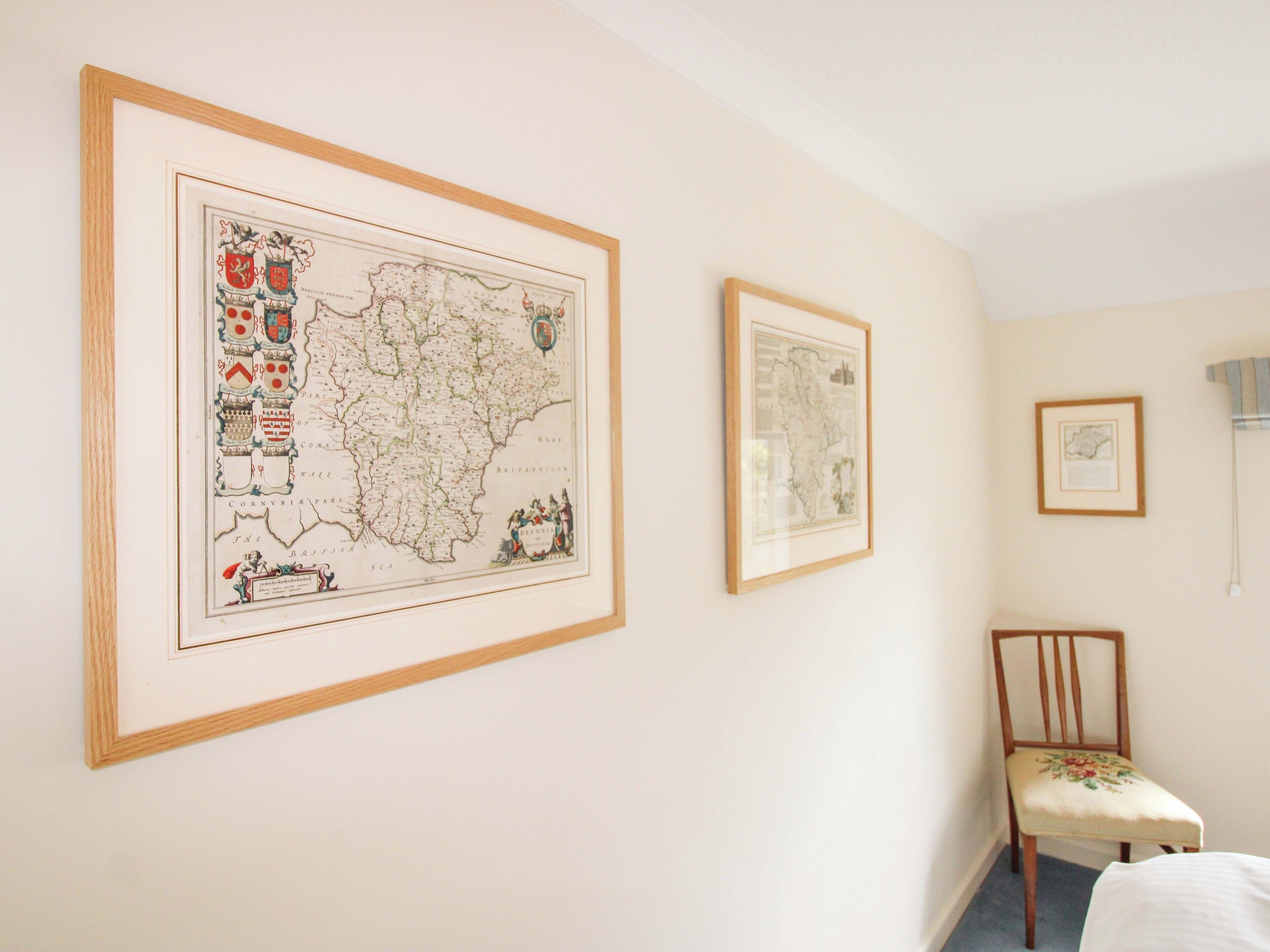 A room with two framed maps on the wall, a wooden chair, and a bed.