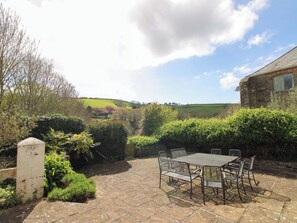 Une terrasse en pierre avec une table et des chaises en métal, entourée de verdure et d’un mur de pierre.