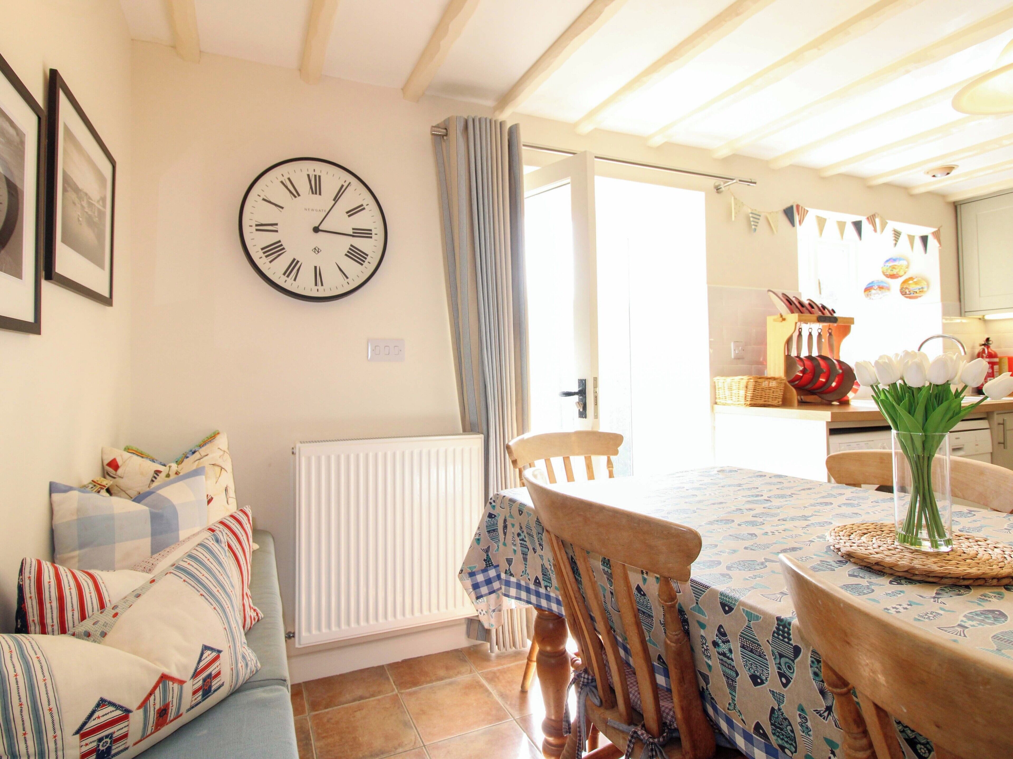 Un comedor con una mesa preparada para dos personas, un reloj en la pared y vista a la cocina.