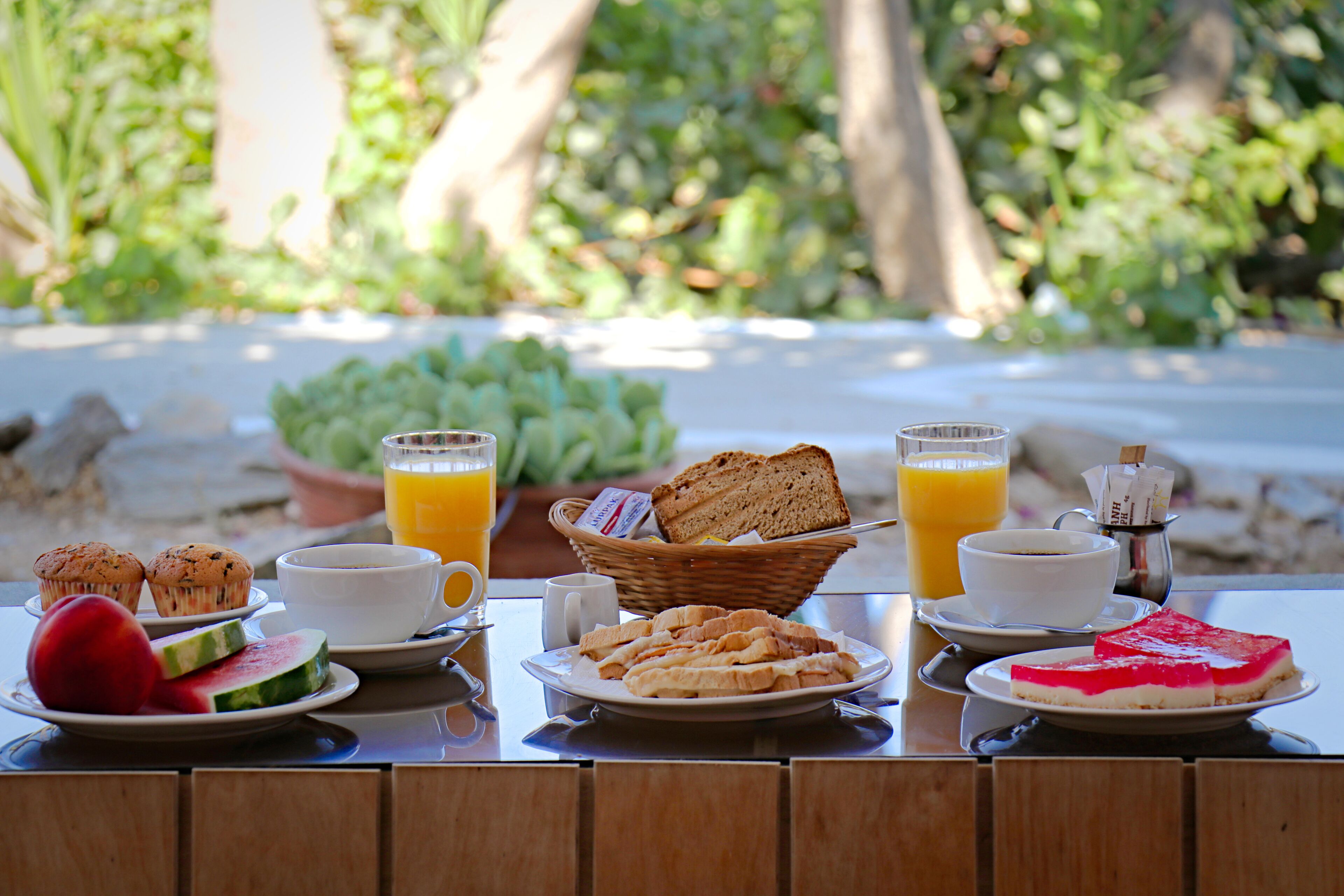A breakfast table with various food items and drinks, set outdoors with greenery in the background.