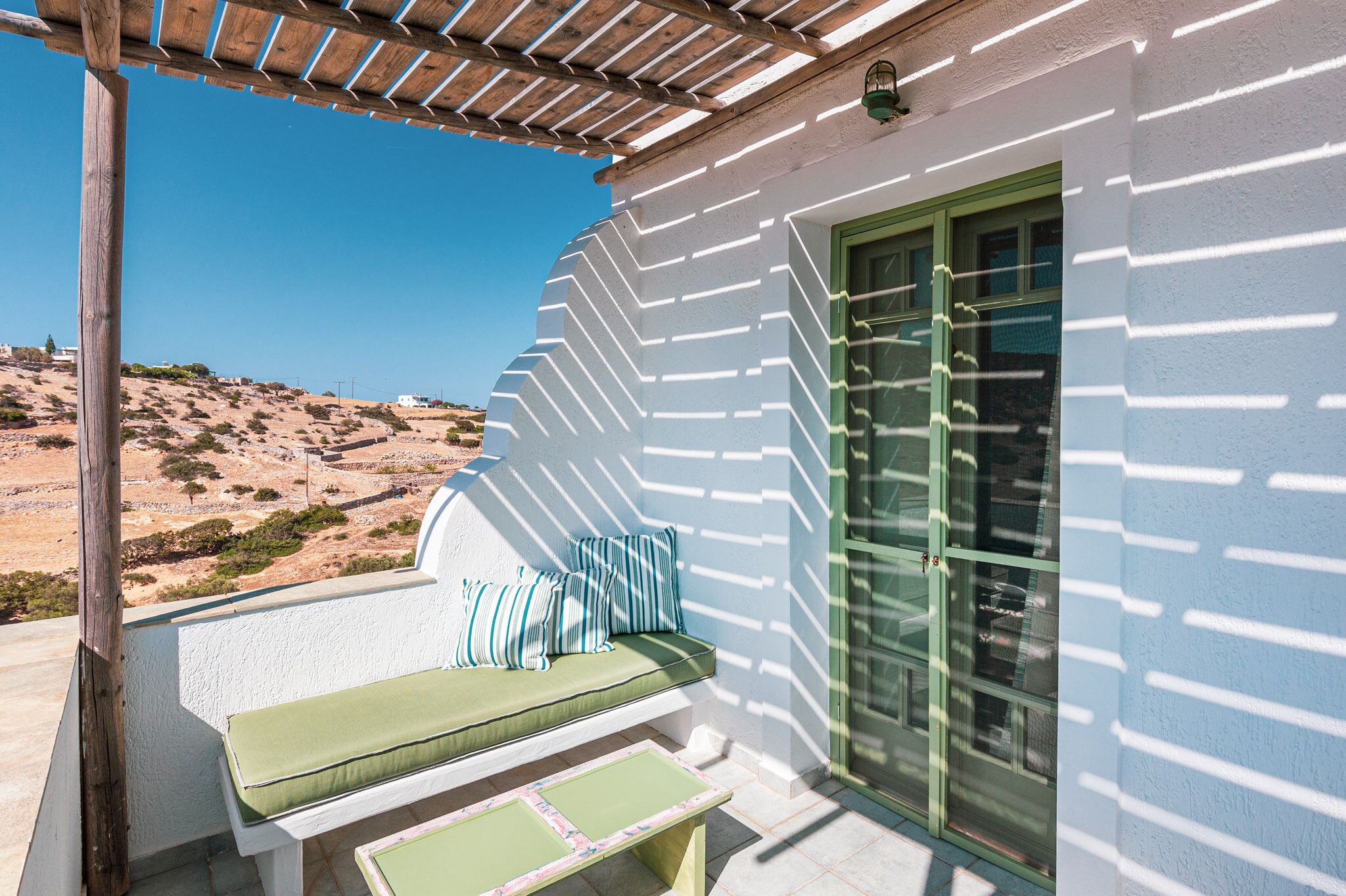A balcony with a wooden pergola, a cushioned seating area with pillows, and a view of a hilly landscape.