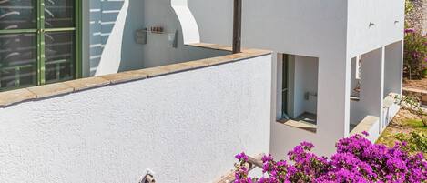 Un edificio moderno blanco con una pérgola, un balcón y flores púrpuras llamativas en primer plano.
