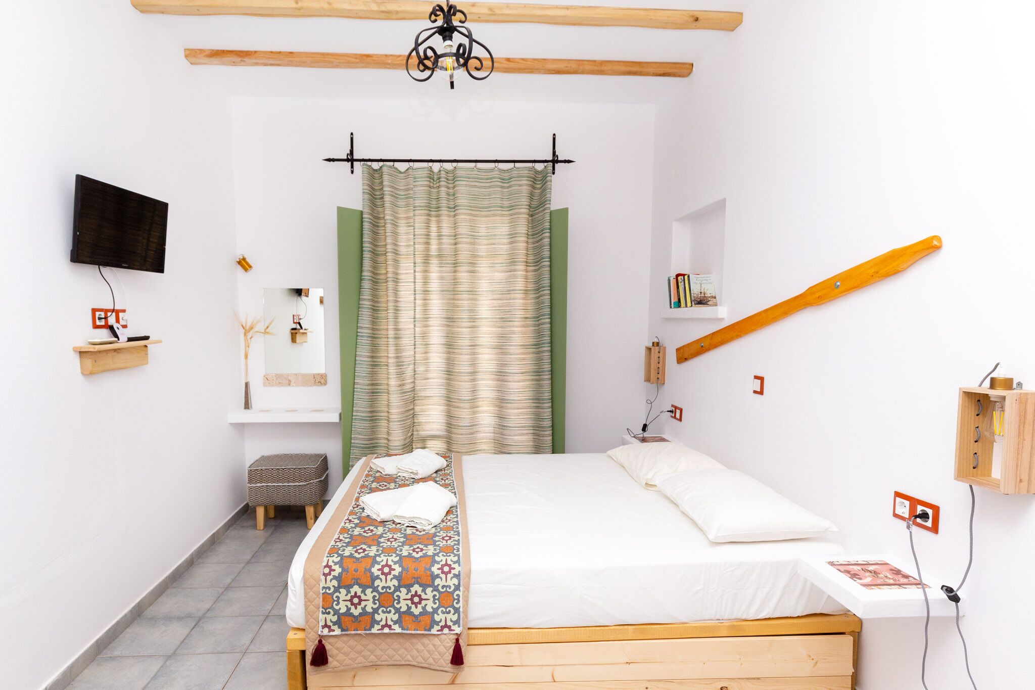 Quarto com cama, televisão, cabeceira de madeira, prateleira pequena e luminária fixada na parede.