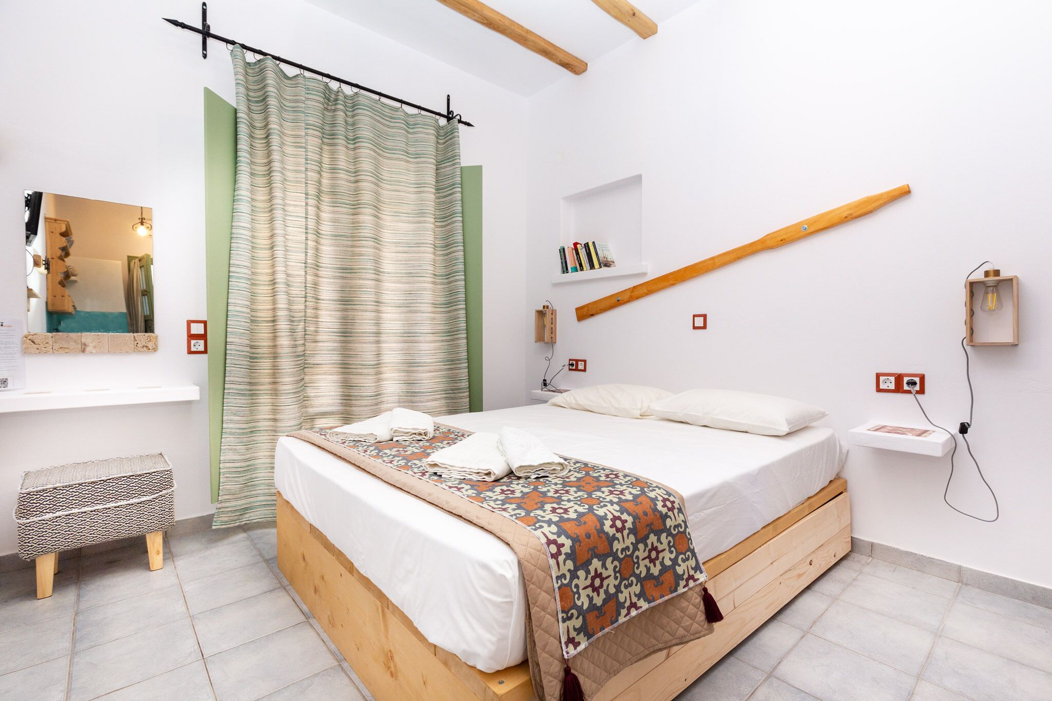 Ein Schlafzimmer mit einem hölzernen Bett, einem grünen Vorhang, einem Nachttisch mit Büchern und einer Wandleuchte.
