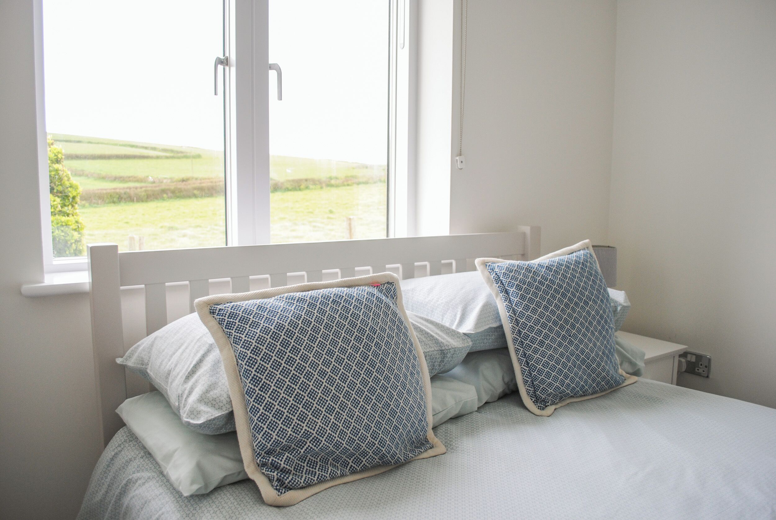 Ein Schlafzimmer mit einem weißen Bett, zwei gemusterten Kissen und einem Fenster mit Blick auf eine grüne Landschaft.
