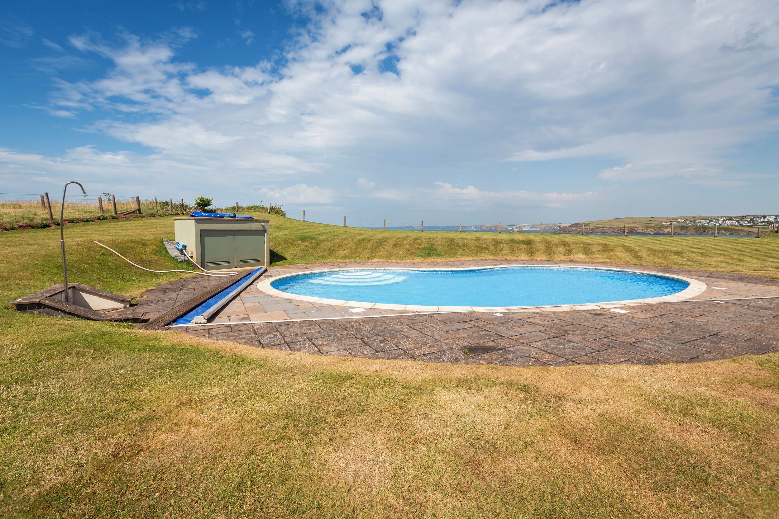 A swimming pool with a surrounding paved area, a small utility shed, and a grassy field with a fence in the background.