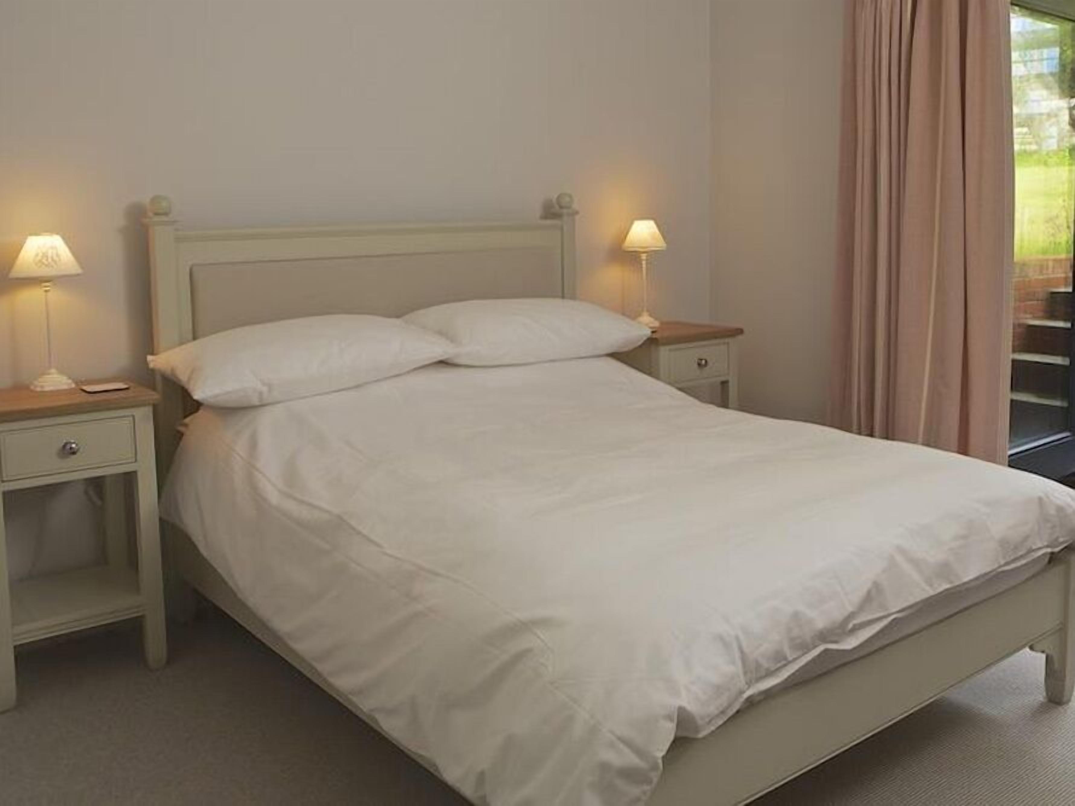 Una cama bien hecha con sábanas blancas, dos mesitas de noche con lámparas y una ventana con cortinas.