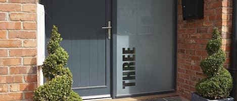 A modern building entrance with a dark grey door, a small potted plant on the left, and another on the right.