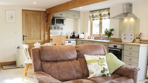 A cozy living room with a brown leather sofa, a wooden door, and a kitchen in the background. - Acorn Cottage (Ludlow)