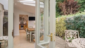 Une porte ouverte donne sur une cuisine moderne avec un coin repas, un téléviseur fixé au mur et un jardin agrémenté d’une table et de chaises en métal blanc.